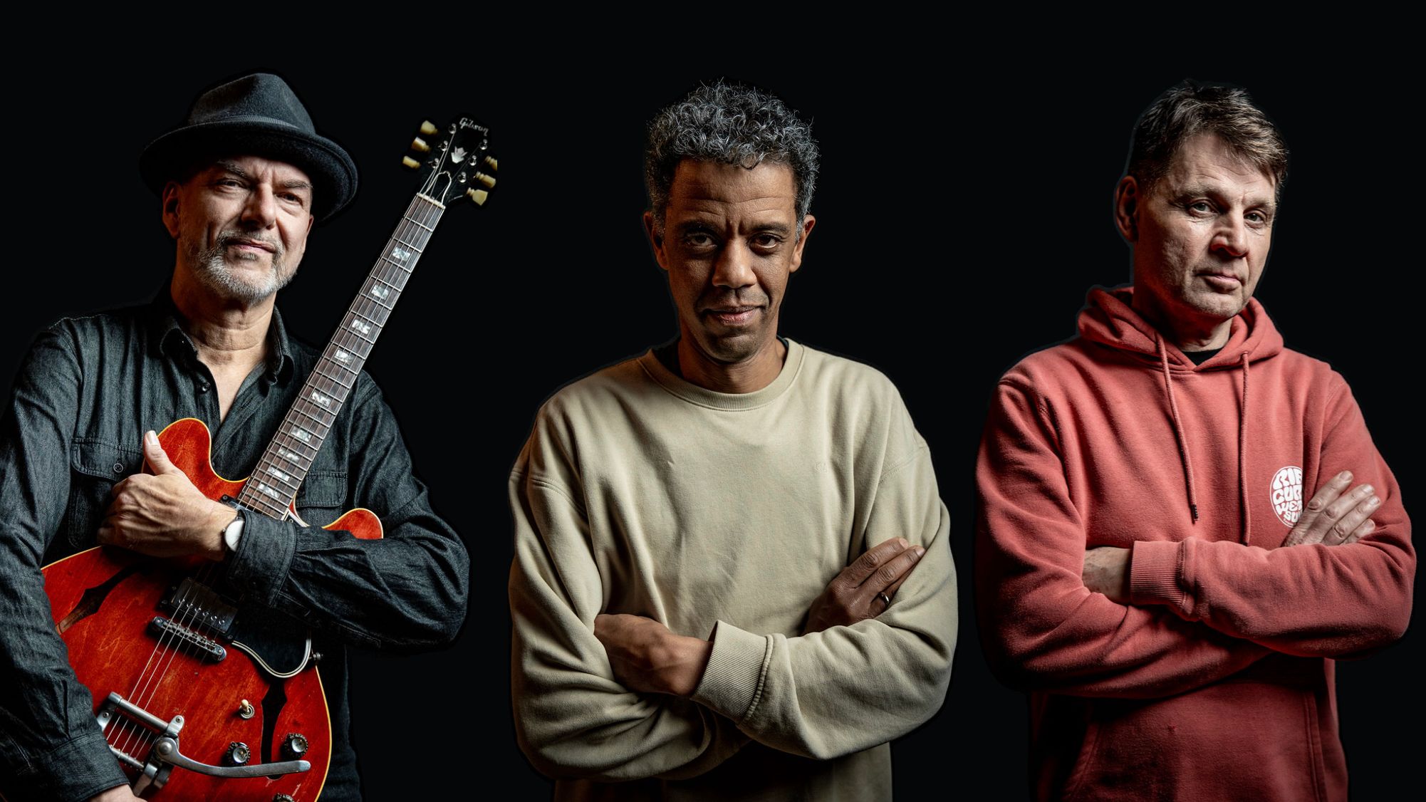 Bandportrait mit drei Männern vor schwarzem Hintergrund, einer mit Gitarre