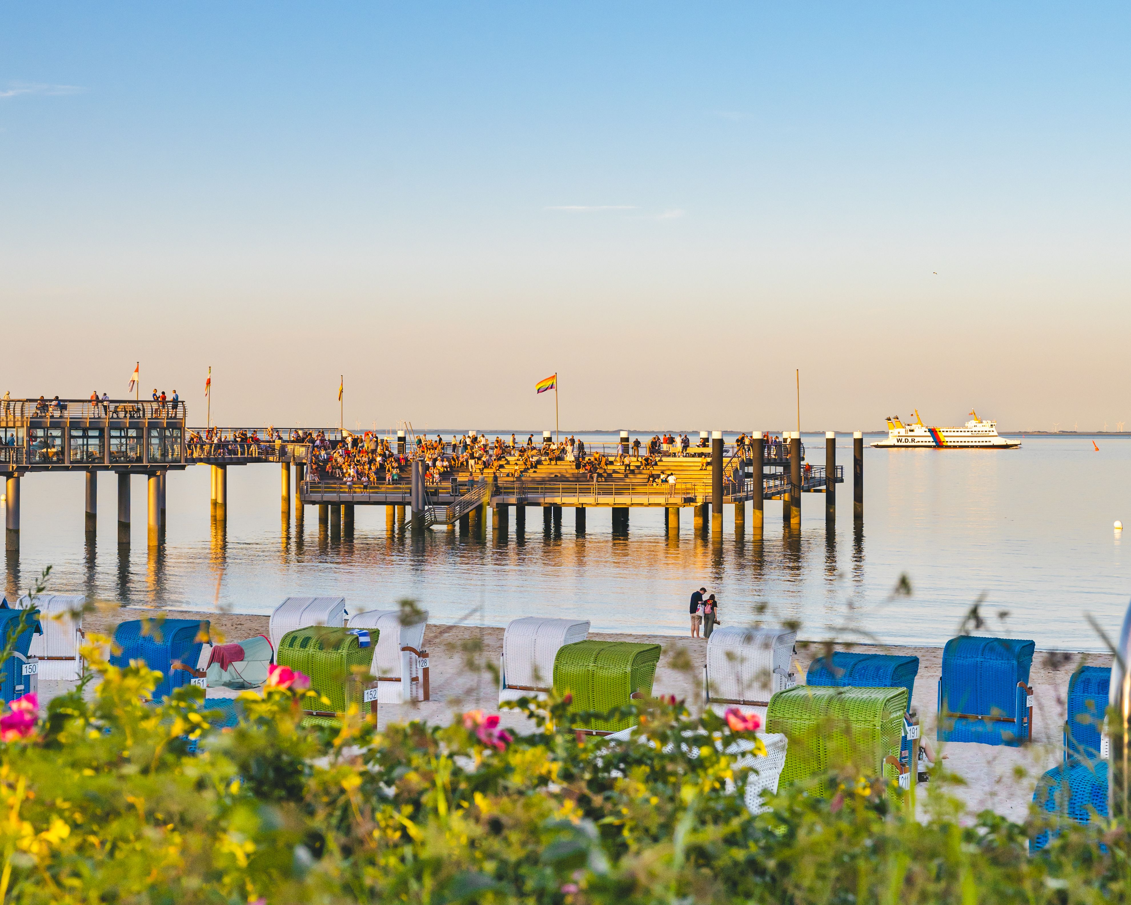 Die Mittelbrücke im Abendlicht mit Publikum beim »Sundowner«-Konzert von »Tone Fish«, im Vordergrund Heckenrosen und Strand mit Strandkörben, im Hintergrund fährt eine Fähre vorbei.