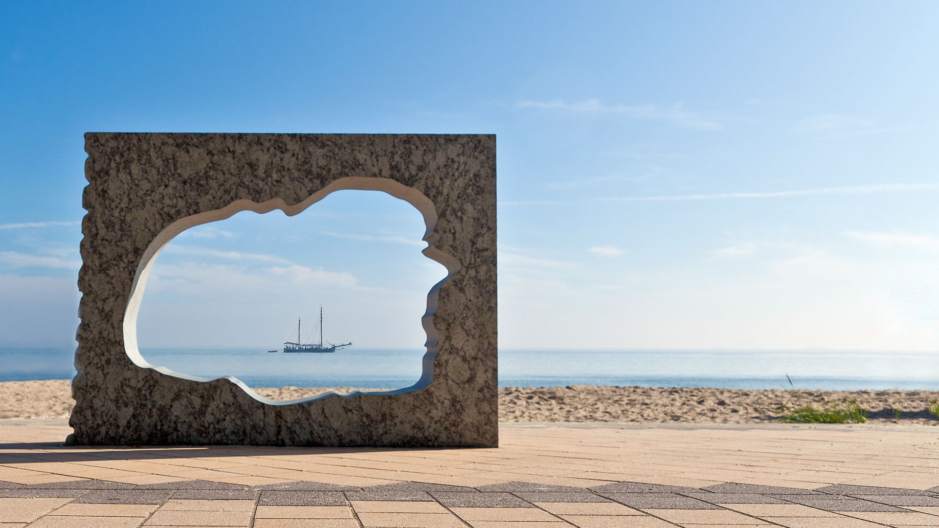 Blick durch eine Steinskulptur mit Inselumriss auf ein Segelschiff an der Strandpromenade in Wyk auf Föhr