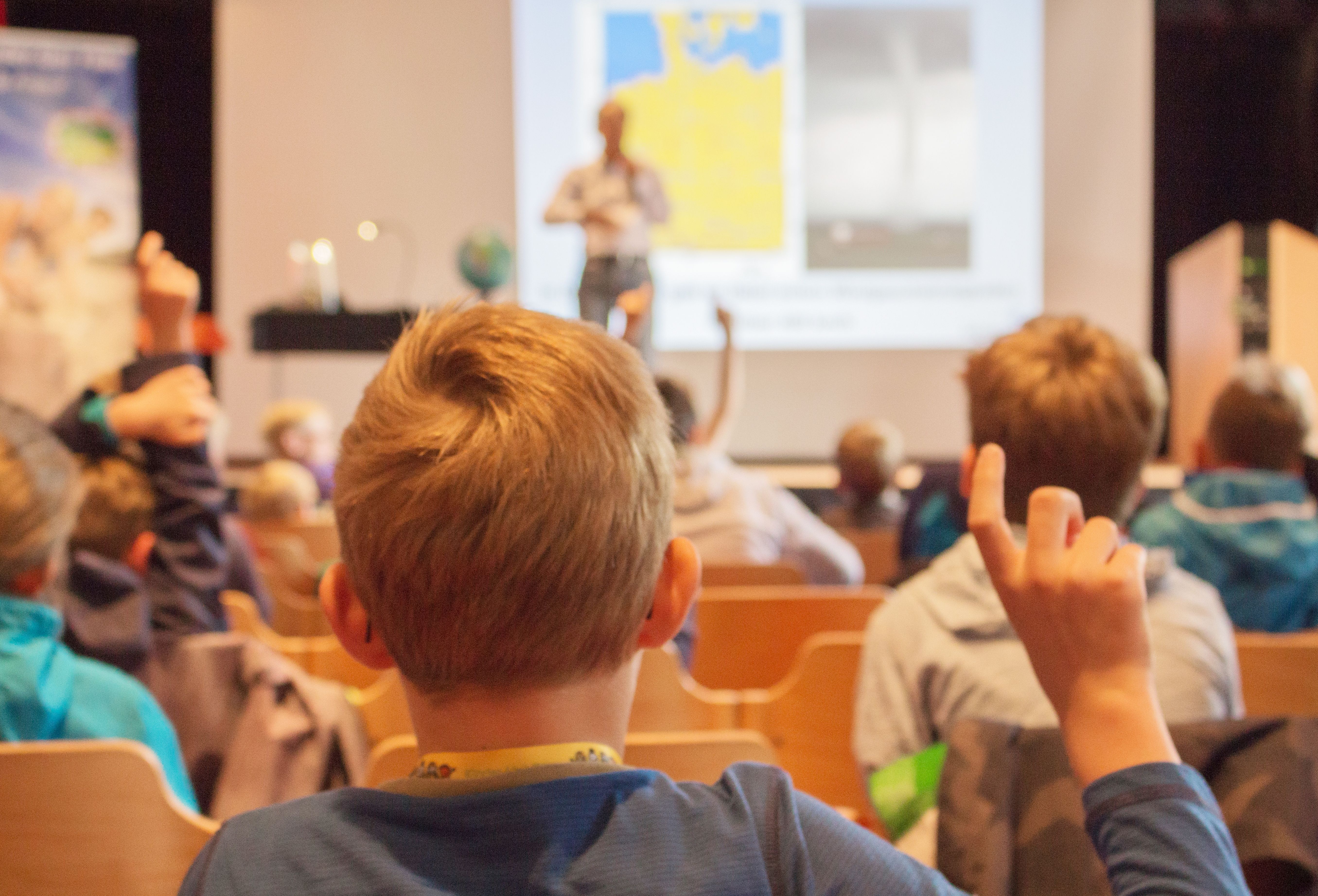 Kind von hinten meldet sich mit erhobenem Zeigefinger im Saal